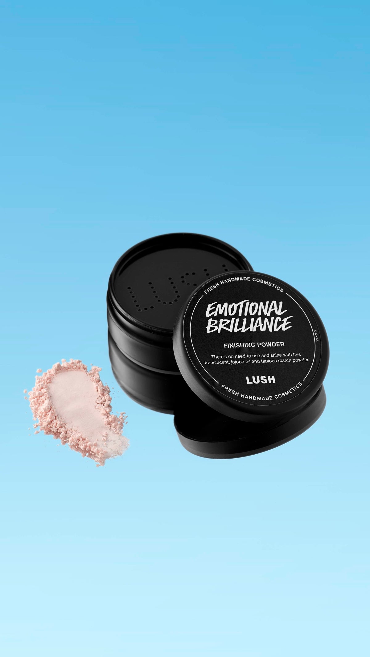 A partially open black pot of Emotional Brilliance ~ Finishing Powder is shown floating against a blue gradient background. A small pile of the pale pink powder is visible next to the pot's base. The black lid has white text identifying the product.