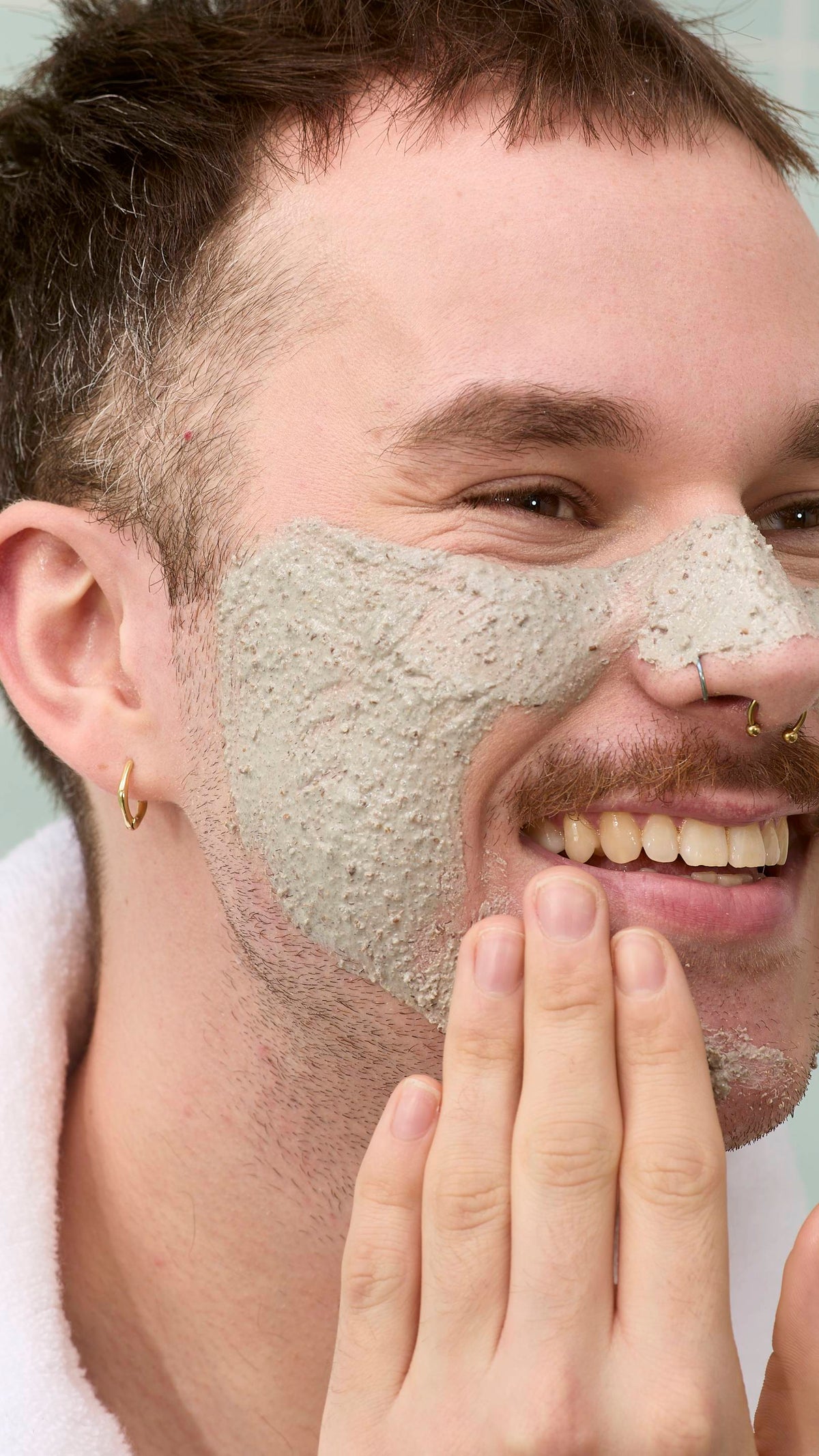 The image shows a model with a moustache and nose piercings smiling while applying a thick, grainy grey "Movis" face mask to their cheek and nose. They are wearing a white fluffy dressing gown against a background of pale green tiles.